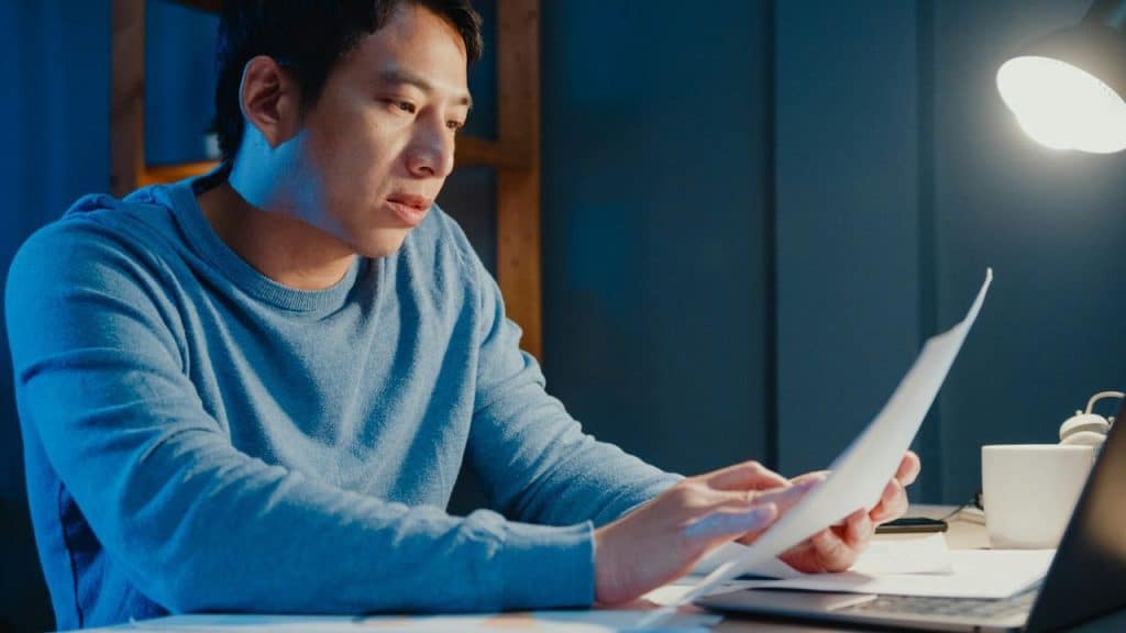 A man in a blue sweater sits at a desk at night, reviewing documents under a lamp.