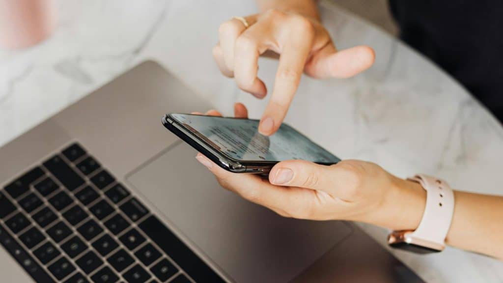 A person using a smartphone above an open laptop.