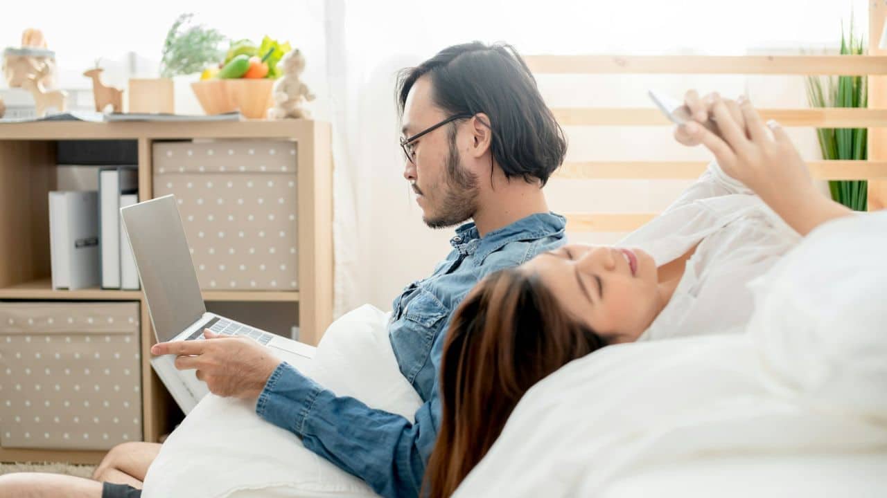 A man uses a laptop while a woman next to him looks at a phone in bed.