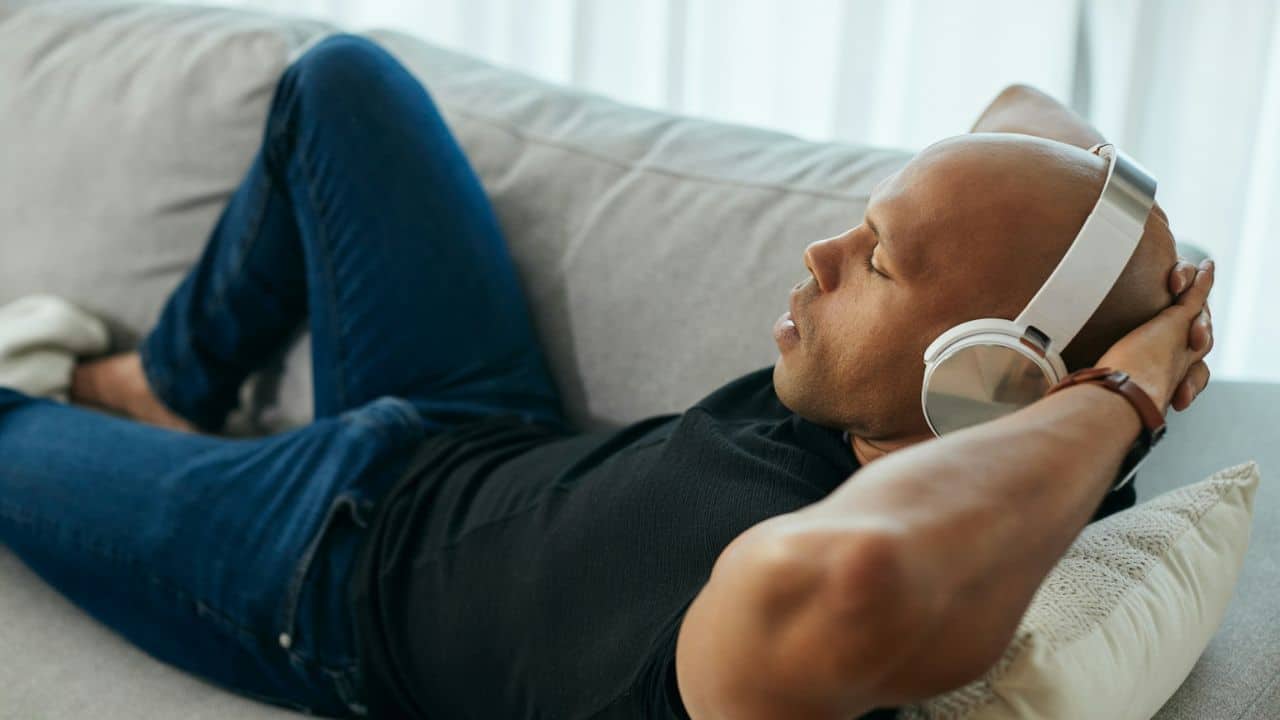 A bald man in jeans and headphones reclines on a couch with hands behind his head.