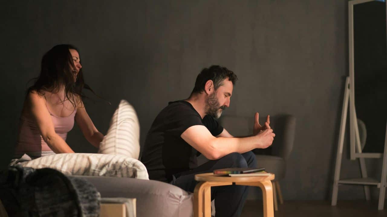 A man sits brooding with his hands clasped as a woman throws a pillow at him.