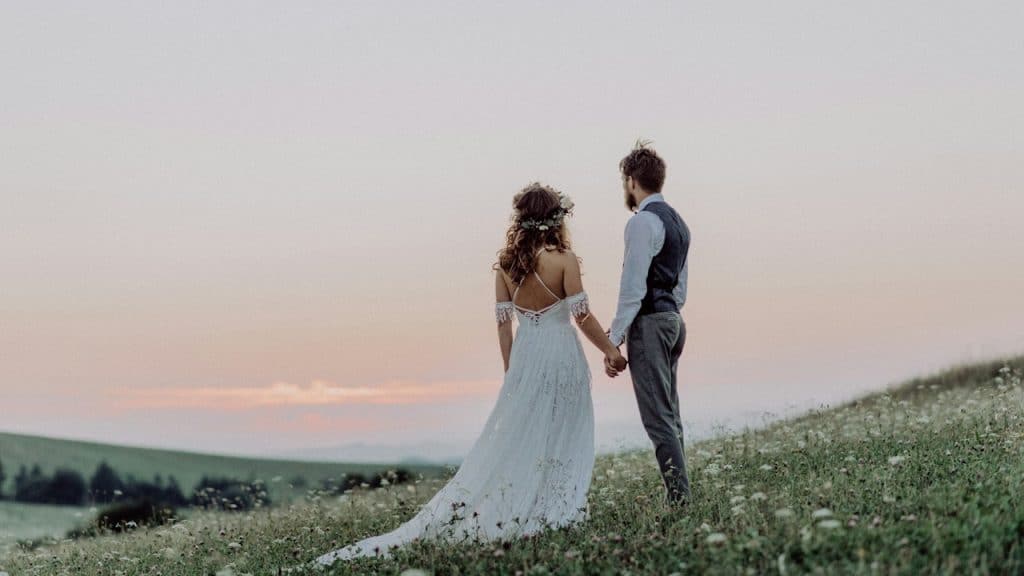 A bride and a groom standing on a hill watching a sunset.