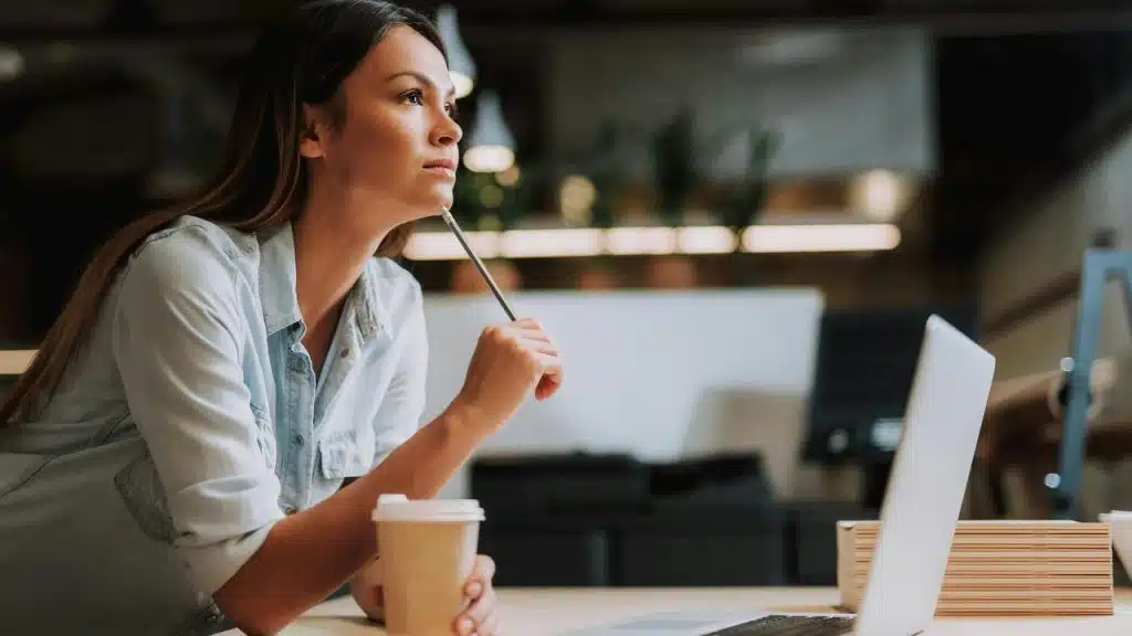 A woman holding a pencil while looking thoughtful at her laptop.