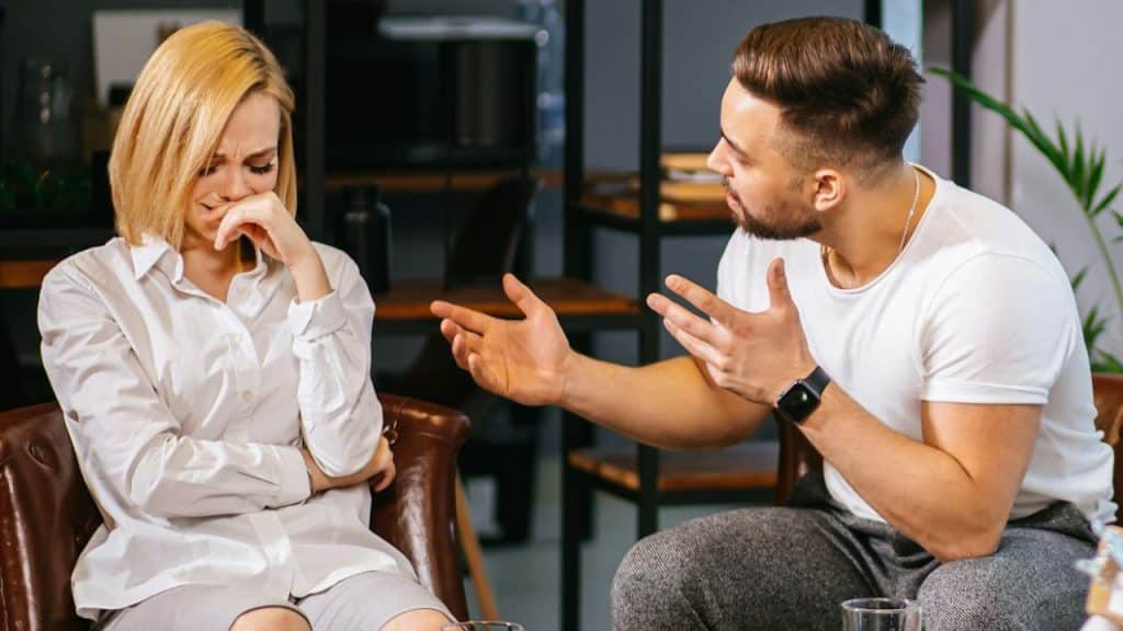 Man gestures strongly toward a woman in a white shirt who is covering her mouth and crying.