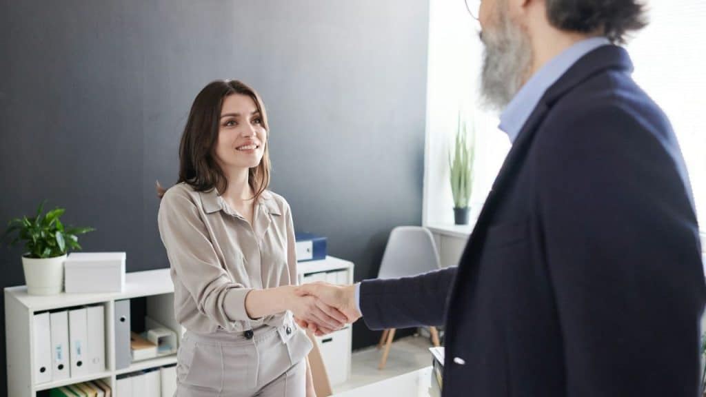 A young woman in a beige shirt smiles while shaking hands with an older bearded man.
