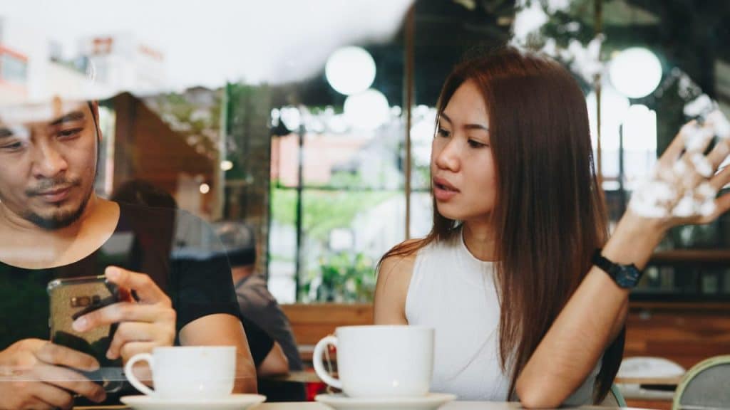 A man checks his phone while a woman looks annoyed and gestures in a café.