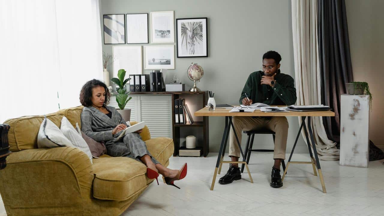 A woman sitting on a couch writing while a man works at a desk nearby.