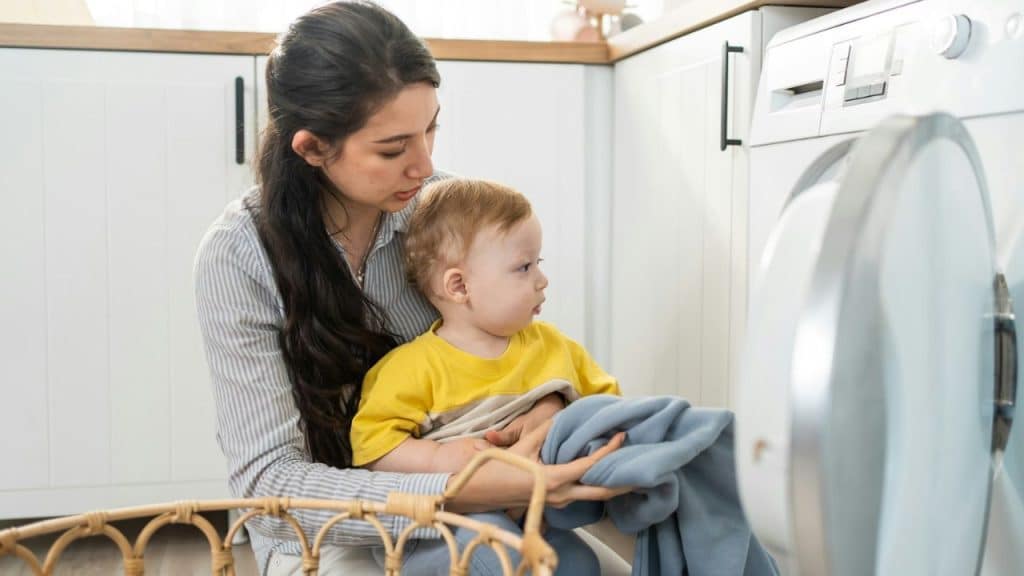 Woman kneeling in laundry room holds a baby while loading a washing machine.