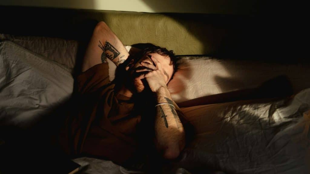 A person with tattooed arms is lying in bed, covering their face with a hand.