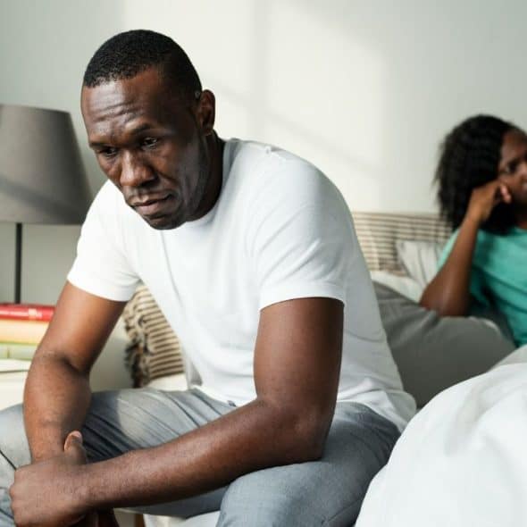 A man sits on the edge of a bed, looking down sadly, with a woman behind him.