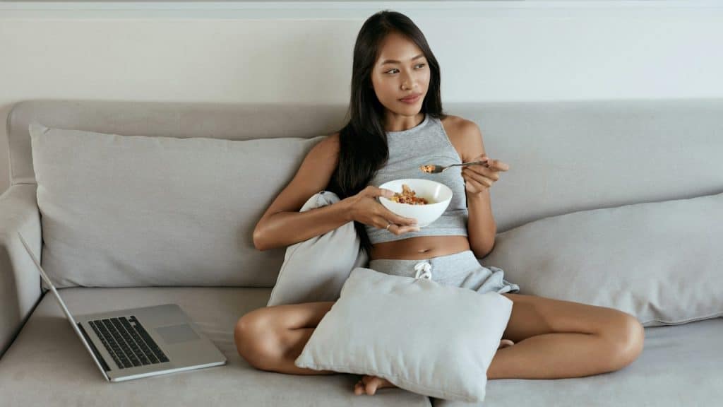 A young woman is sitting cross-legged on a gray sofa eating from a bowl with a spoon, next to an open laptop.