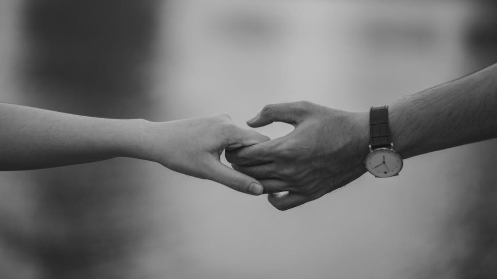 A black and white close-up image of a man and woman holding hands with the man wearing a wristwatch.