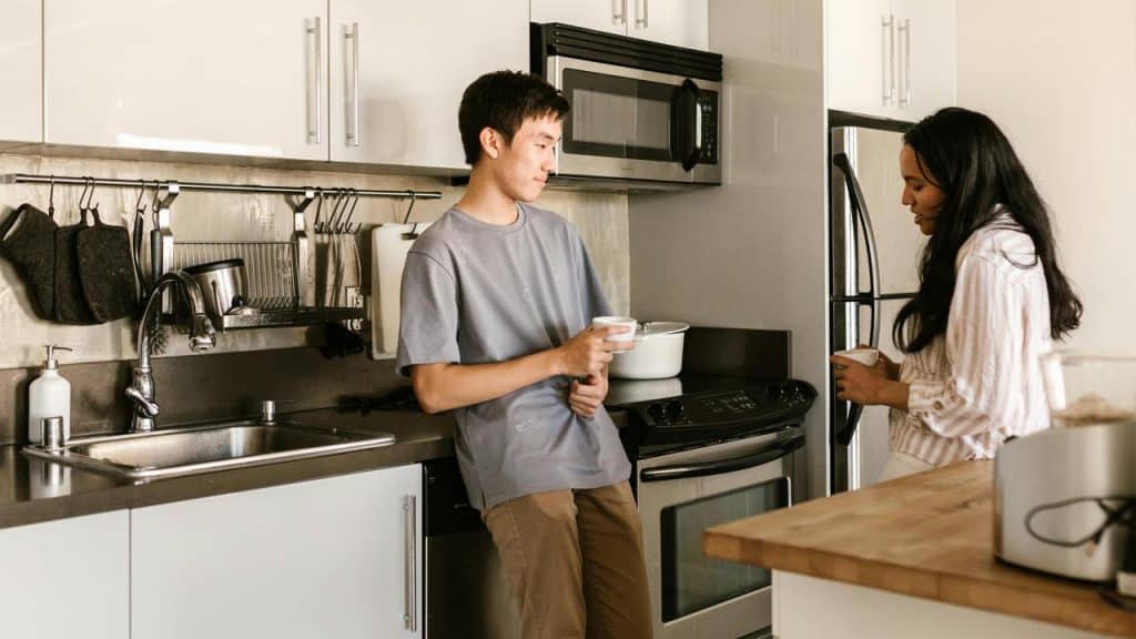 A man and woman chatting in a kitchen while holding mugs.