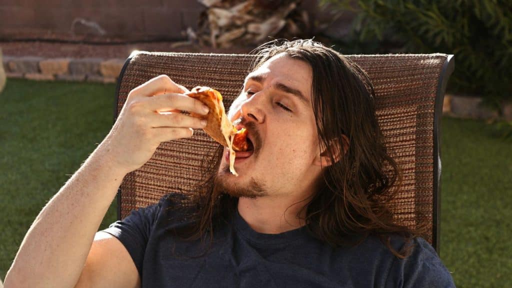 A man with long hair and a beard is eating a slice of pizza outdoors.