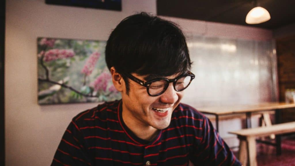 Smiling man with dark hair and glasses sitting at a table indoors.