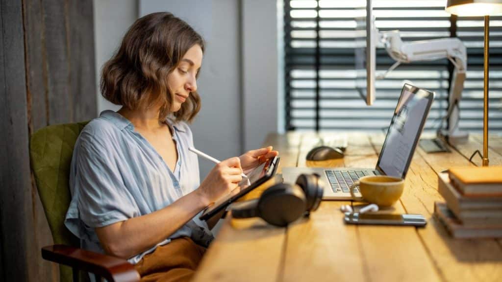 Woman with short brown hair drawing on a tablet at a wooden desk.