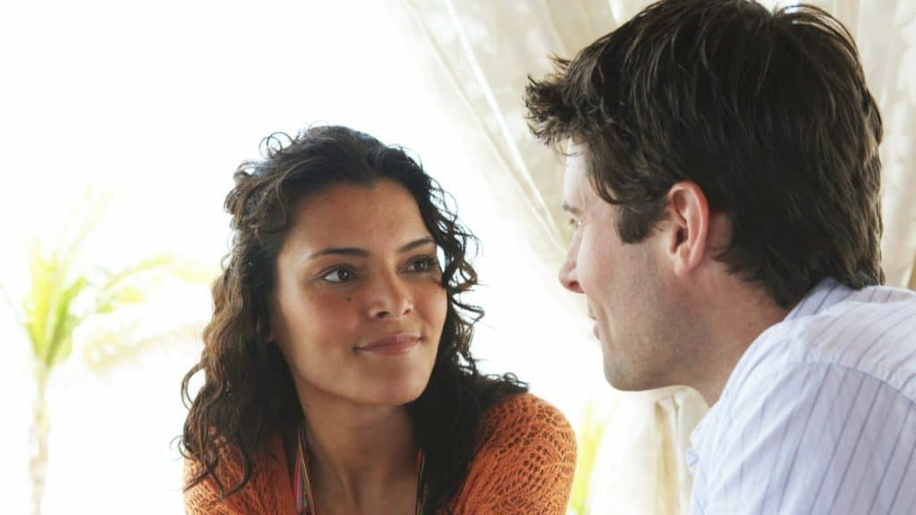 A woman with dark curly hair smiling and looking at a man in a white shirt outdoors.