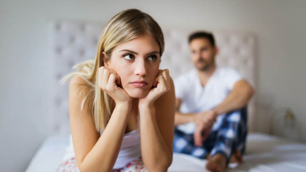 Blonde woman sitting on a bed with hands on cheeks looking away, man is blurred in background.