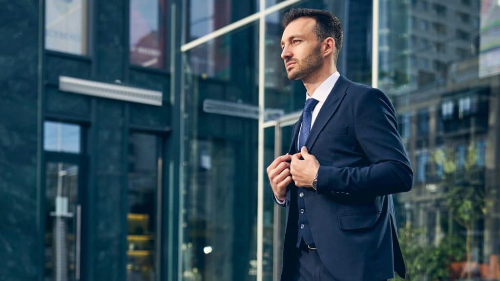 Man in a navy blue suit and tie adjusting his jacket in a modern city setting.