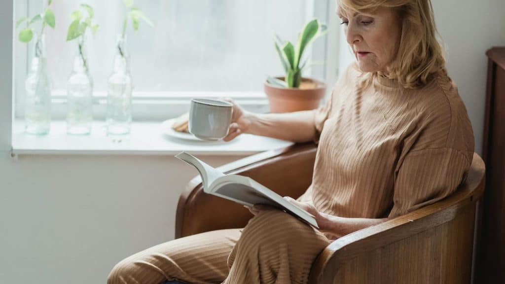 An older woman sitting by a window reading a book and holding a cup.