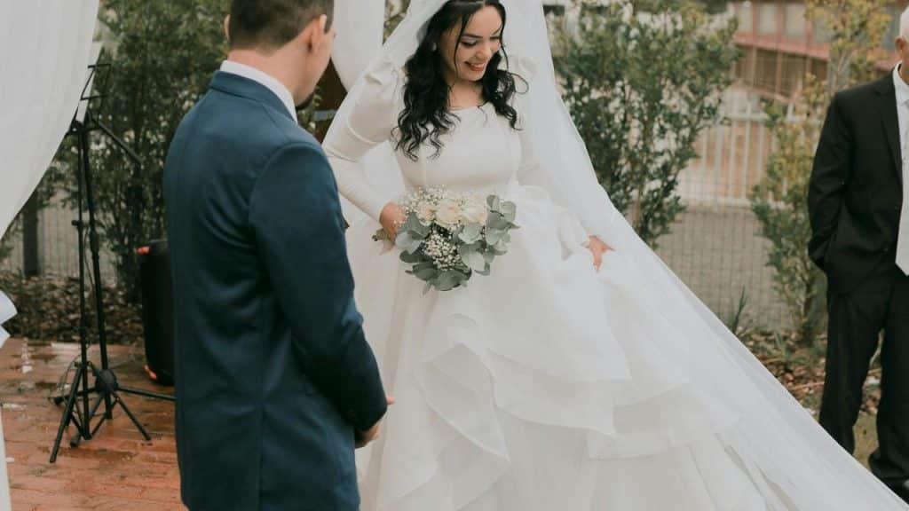 A smiling bride holds a bouquet while looking at the groom outdoors.