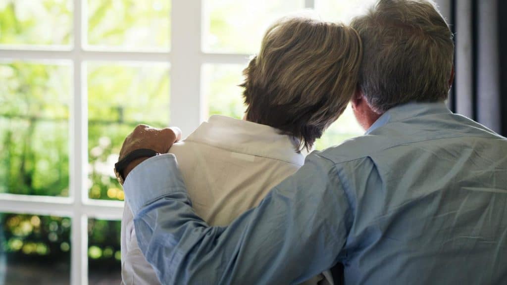 An older couple sits together, embracing while looking out a bright window.