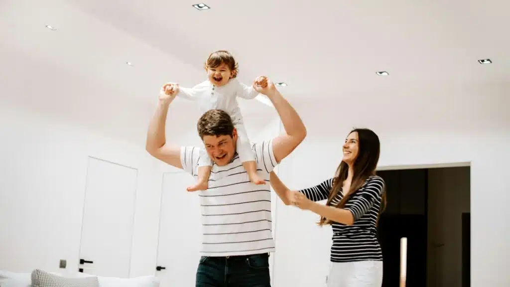 A man holding a little girl on his shoulders and his wife is smiling.