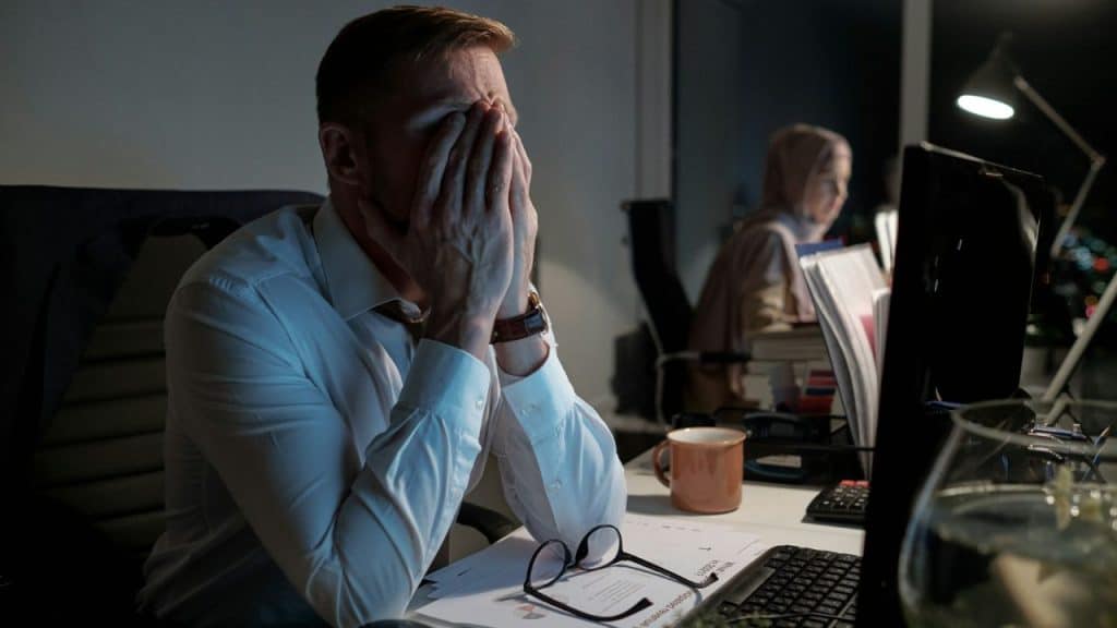A tired man in a white shirt rubs his eyes while sitting at a desk late at night.