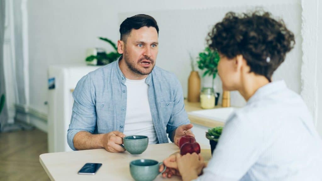 A surprised man sits at a table, holding a mug, talking to a woman across from him.