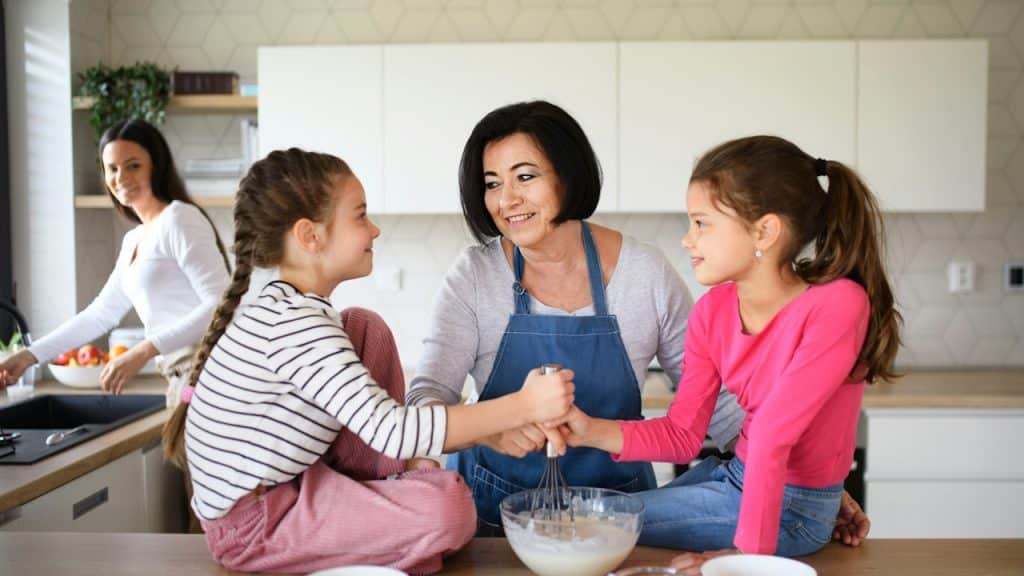 Happy little girls with their mother and grandmother making pancake mixture at home.