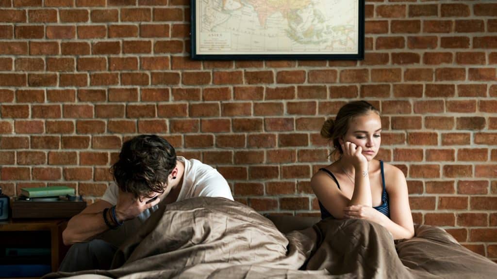 A couple sitting on a bed after a fight, with both people looking unhappy and facing away from each other.