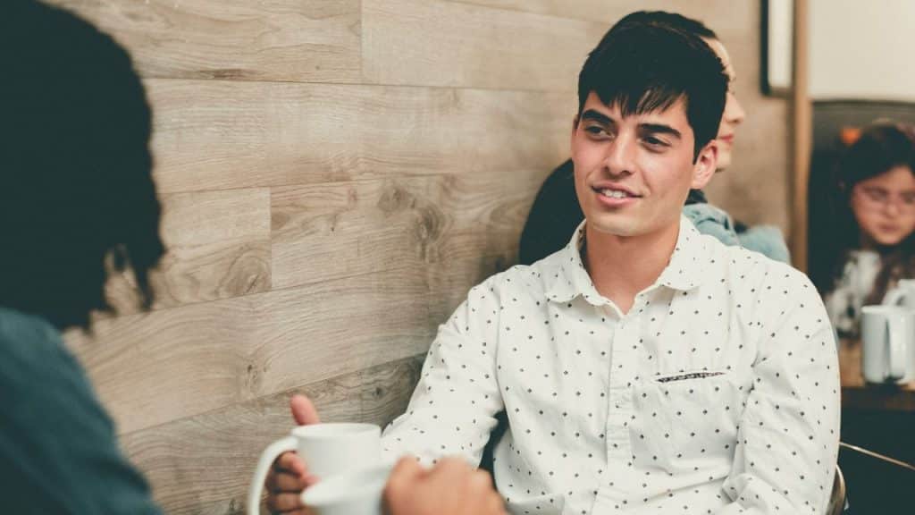 A smiling young man in a white collared shirt sits opposite another person.