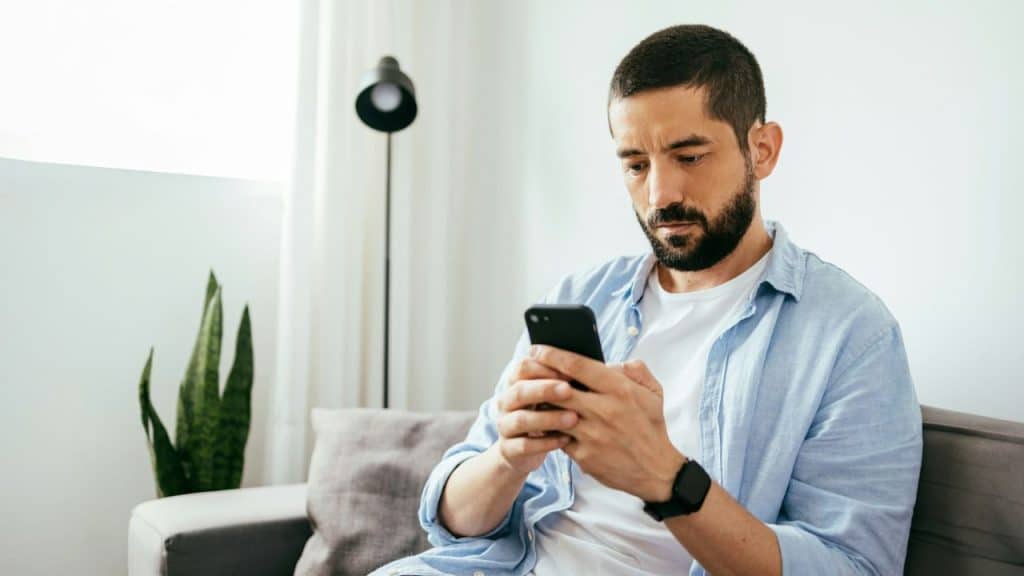 A focused, bearded man sits on a couch, looking at his smartphone.