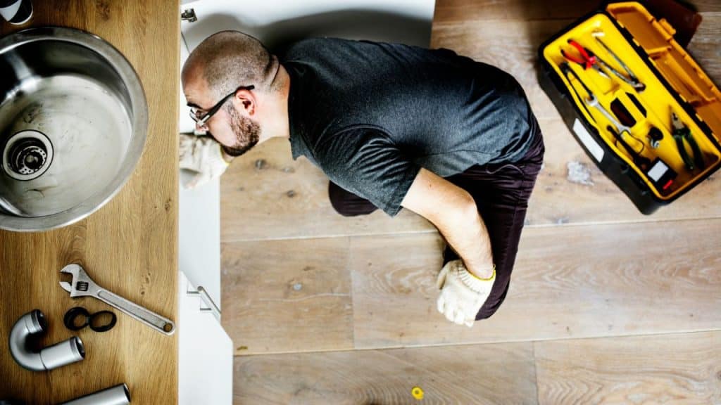 An overhead view of a man on a wooden floor fixing a sink with a wrench and a yellow toolbox nearby.