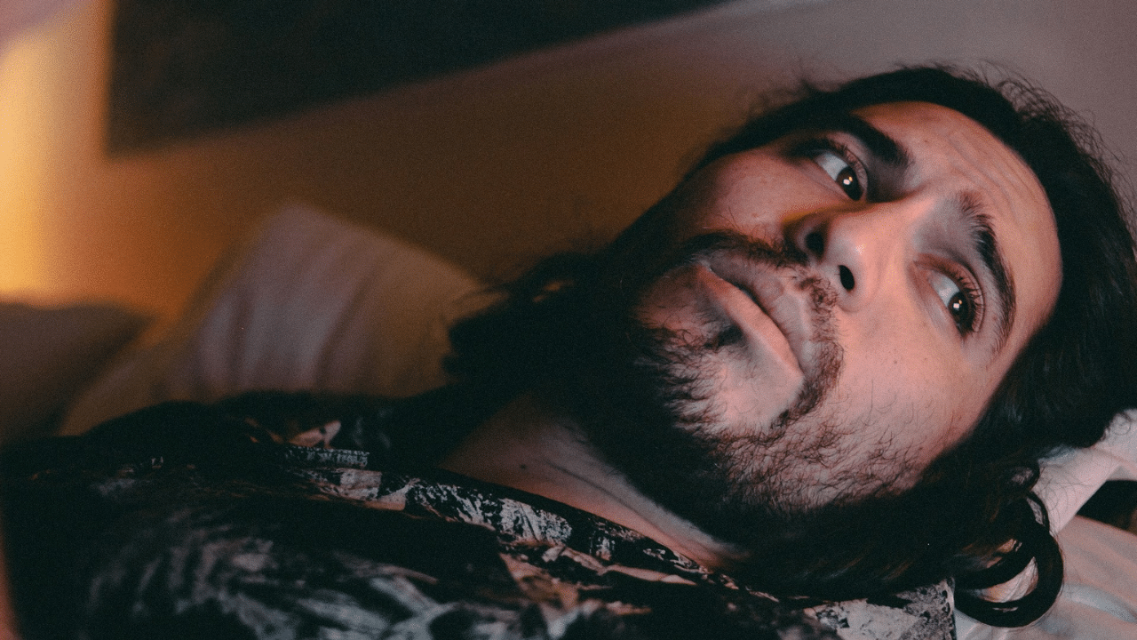 A bearded man with long hair is lying down, looking up with his eyes wide open.