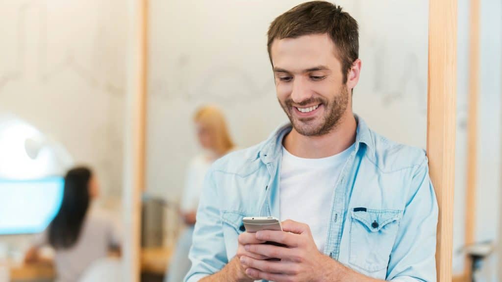 A handsome man in a light blue denim shirt and white T-shirt smiles while looking down at and texting on his smartphone.