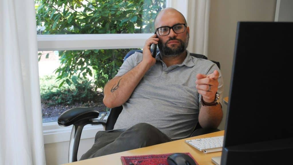 Bald man with a beard and glasses sitting at a desk on a phone call, pointing forward.
