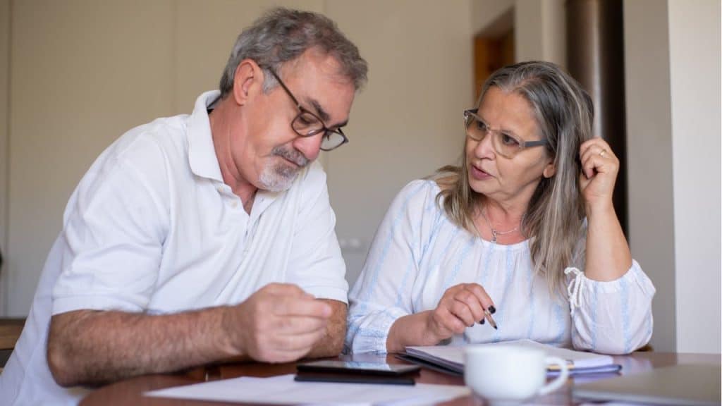 Two middle-aged individuals wearing glasses sit at a table looking at papers, appearing to discuss finances or documents.