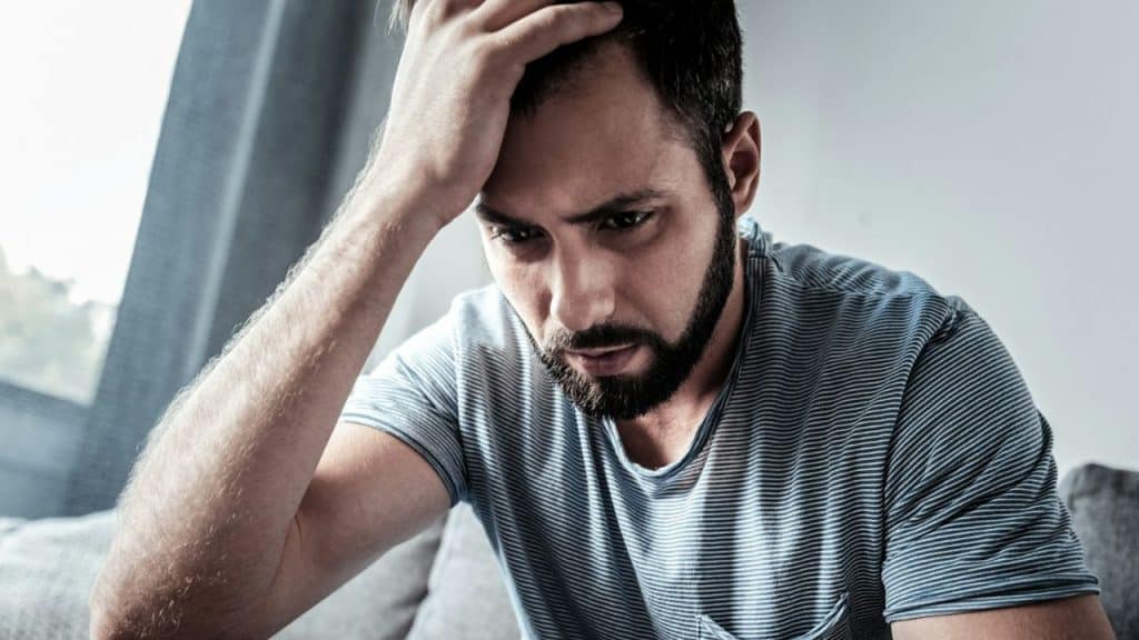 Man with a beard in a striped shirt sitting, holding his hand to his head with a troubled expression.