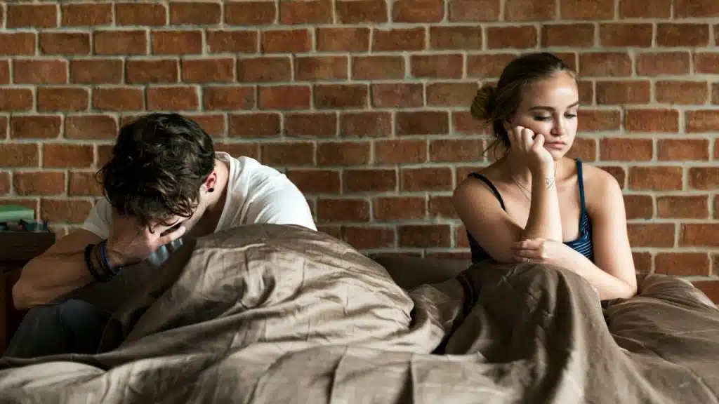 Distressed man and woman sitting apart on a bed with a brick wall behind them.