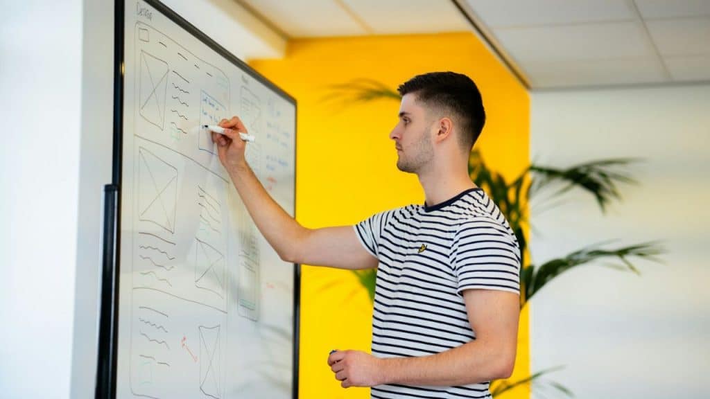 Man in a striped shirt drawing diagrams on a large whiteboard in an office.