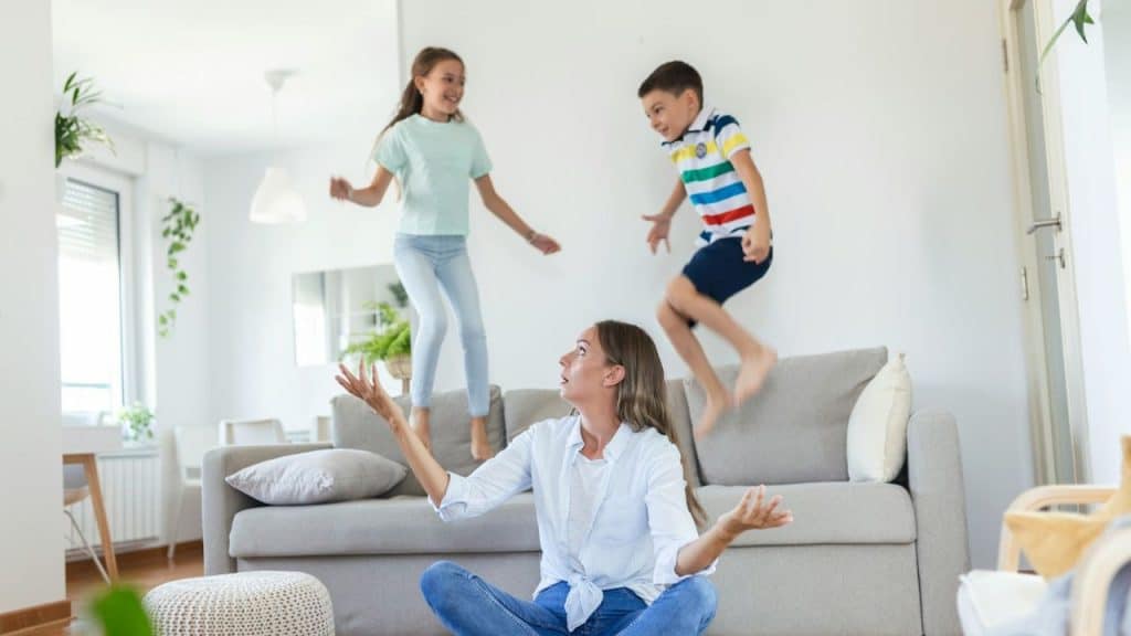 Woman on the floor looking up in surprise as two children jump on the couch.