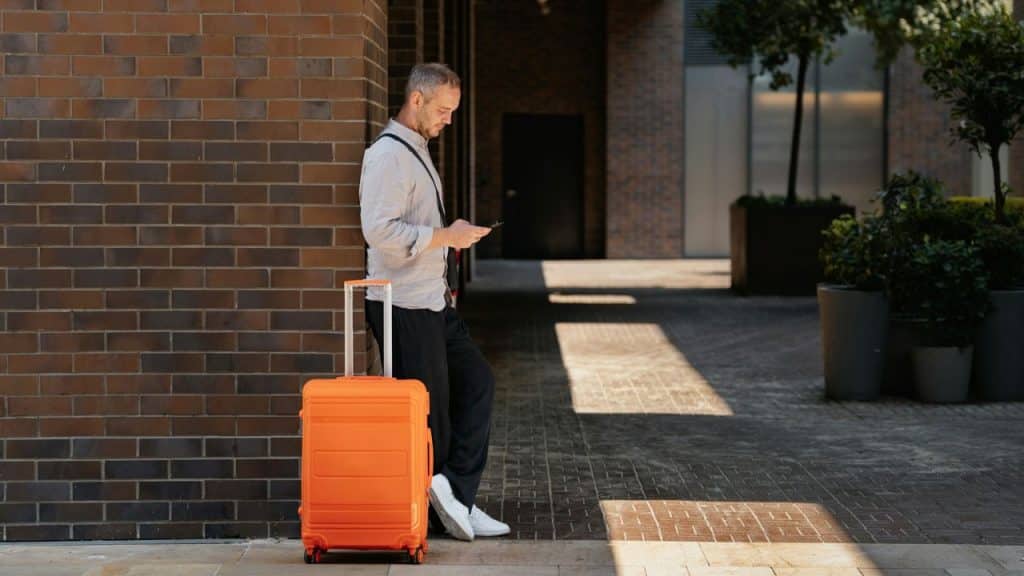 Man leaning against a brick wall with an orange suitcase, looking at his phone.