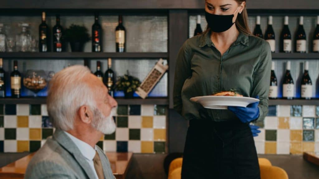 Waitress wearing a face mask and gloves serving a dish to an elderly man in a restaurant.