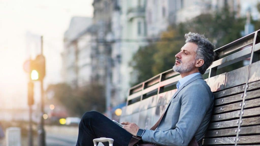 A relaxed, middle-aged man with eyes closed sits on a wooden bench outdoors.