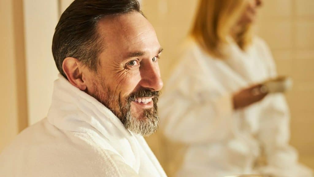 Middle-aged man in a white robe smiling, with a blurred woman in the background.