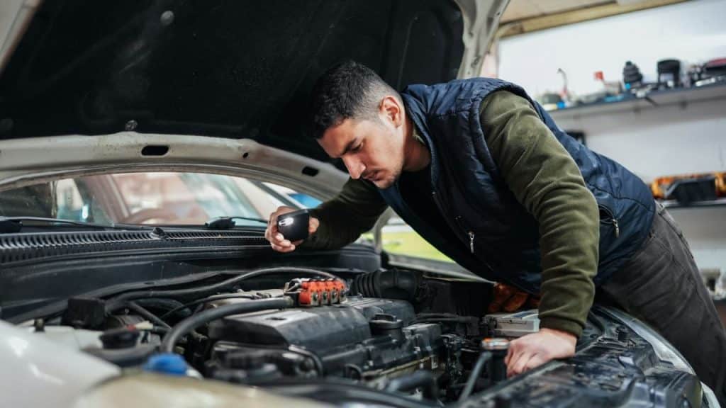 Man wearing a vest inspecting a car engine with a small light in a garage.