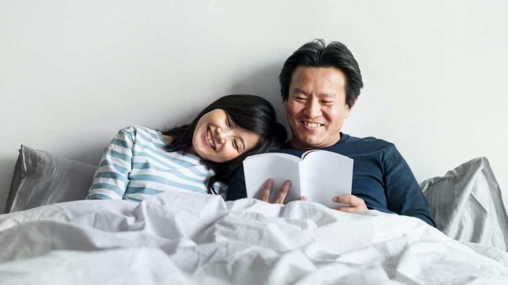 Smiling couple reading a book together in bed with a white wall as the background.