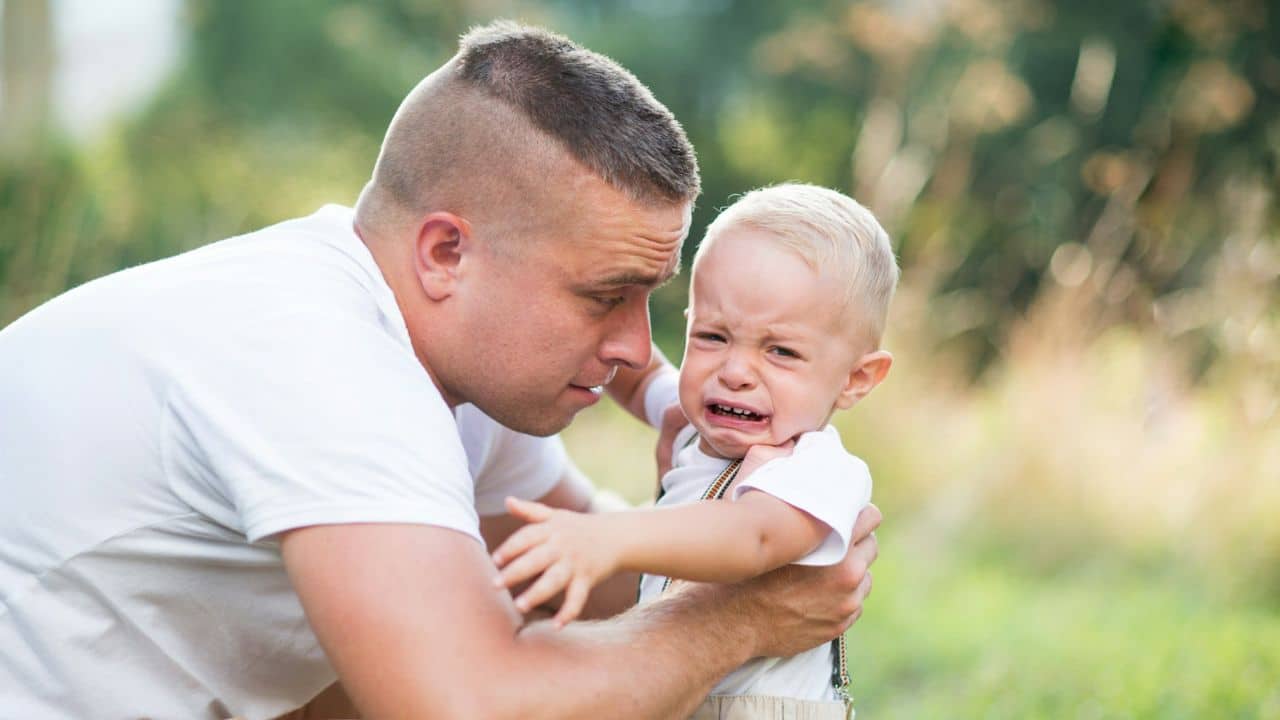 Man holding a young blonde boy who is crying outdoors in a grassy area.