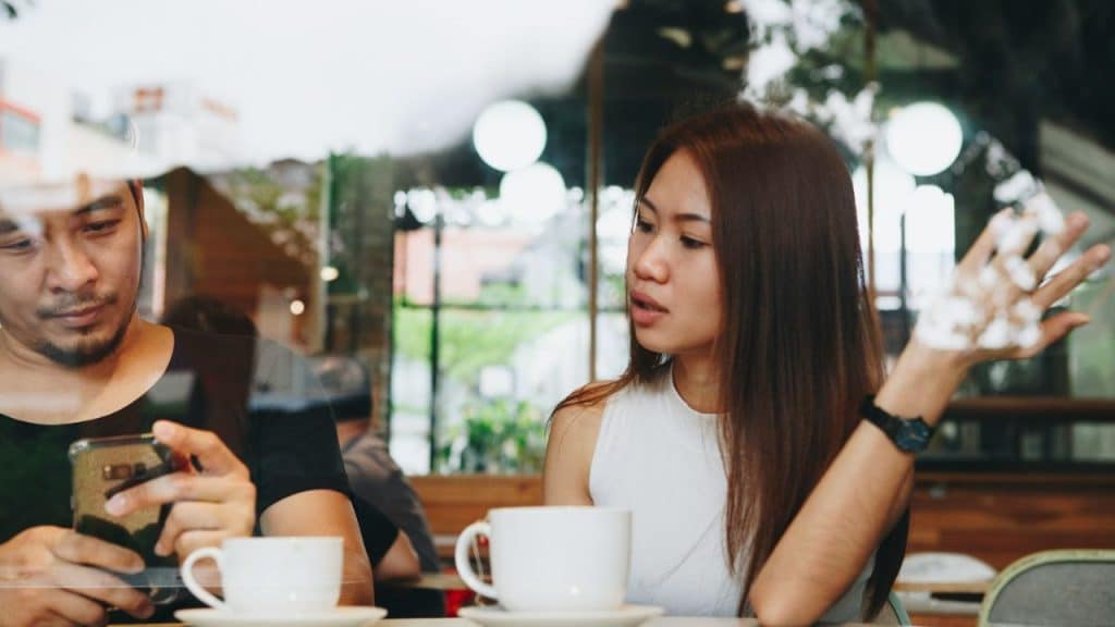 Woman talking and gesturing to a man distracted by his phone in a cafe.